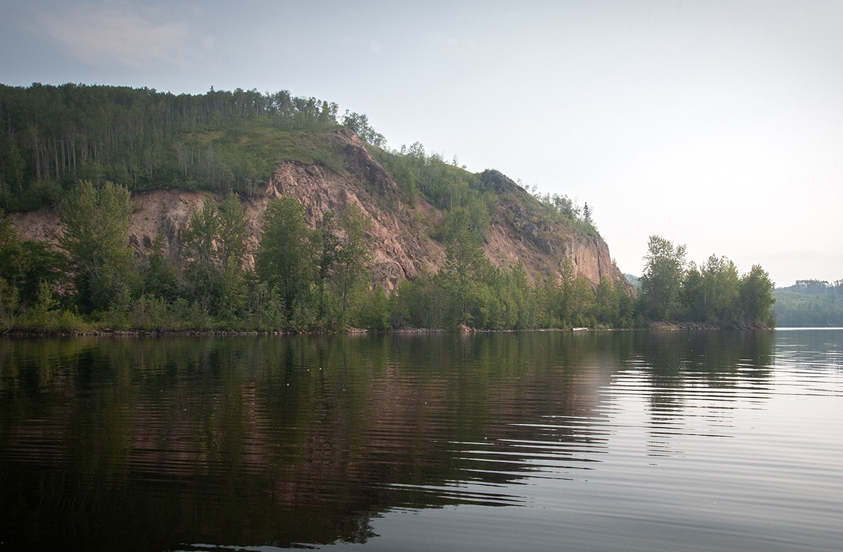A rocky cliff with forest at the top and base of it. There is blue sky behind and a water body in the foreground.