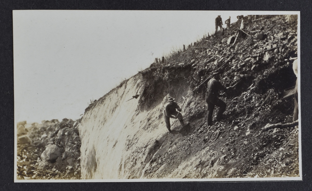 A black and white photo shows men standing on a steep hillside and using tools to remove rock.