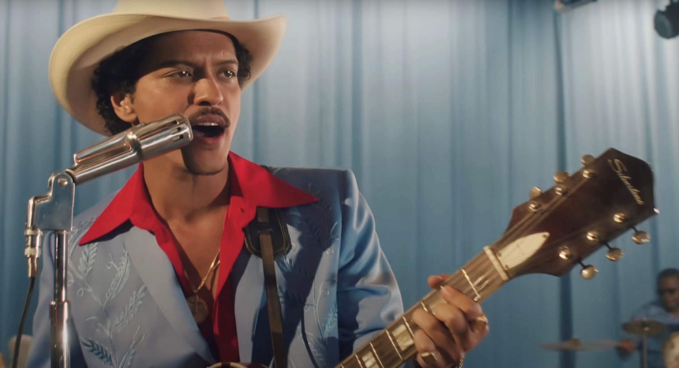 Bruno Mars has dark curly hair, a moustache and a medium skin tone. He&rsquo;s wearing a cowboy hat, and a patterned silver blaze over a pink shirt. He is playing a guitar and singing into a microphone onstage with silver curtains in the background.
