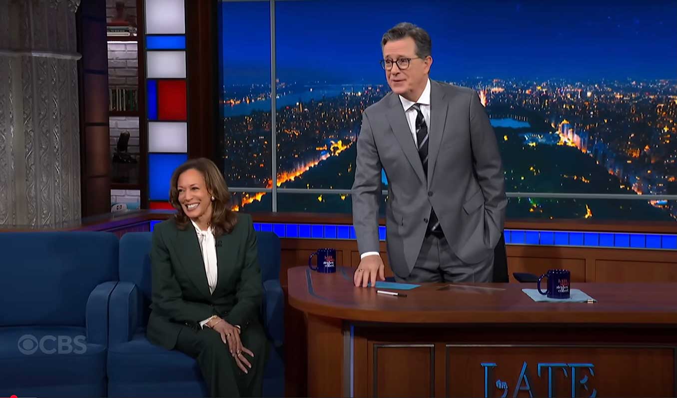 Kamala Harris, left, is seated on a navy chair. She is wearing a hunter green suit over a white shift. She has a medium skin tone and shoulder-length brown hair. She is smiling at the audience offstage. To her right, Stephen Colbert is standing behind a wooden desk. He has a pale skin tone and wavy greying hair with glasses. He&rsquo;s wearing a grey suit over a white shirt and dark tie, and a digital projection of a city at night is behind him.