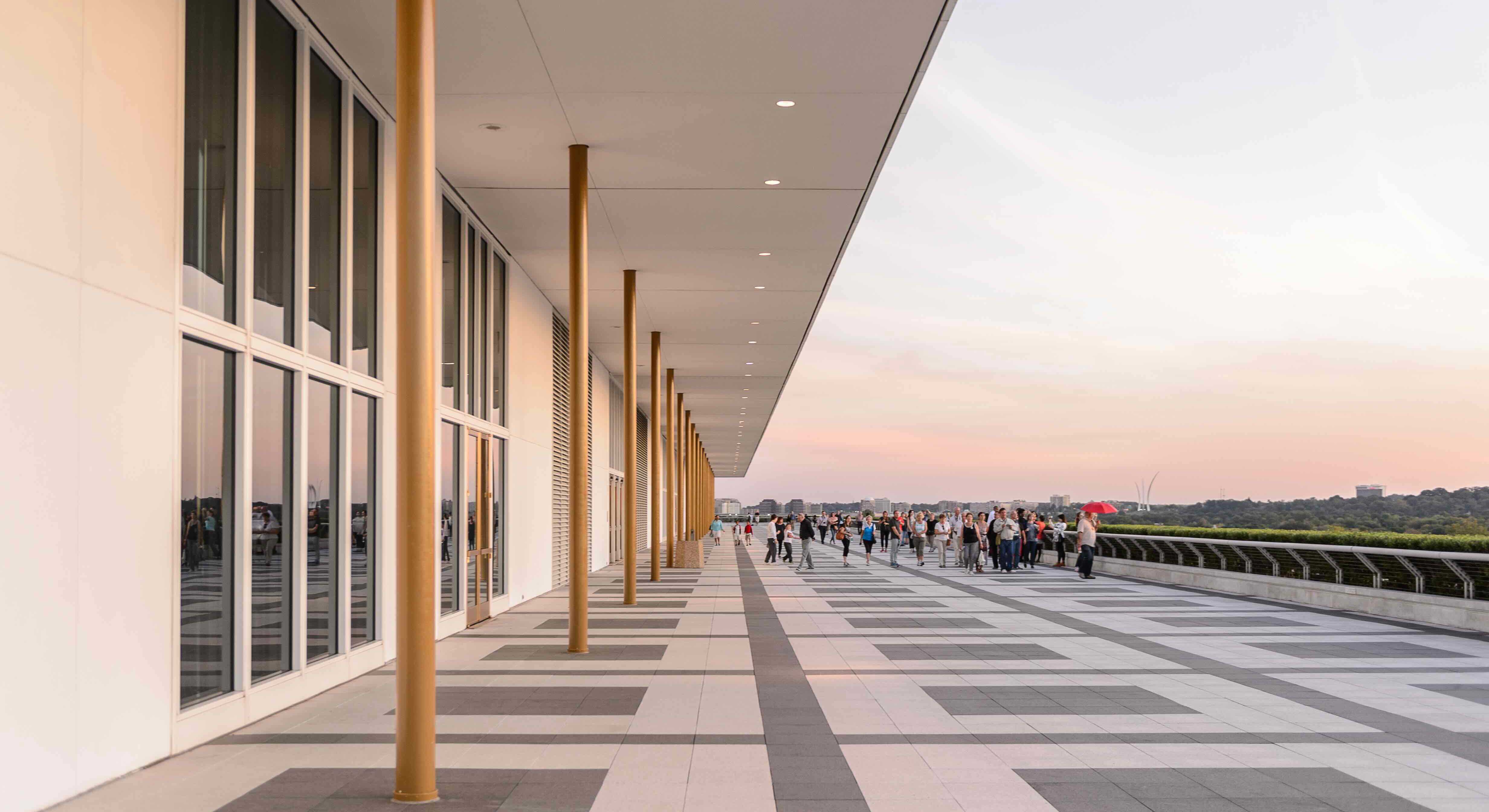 The John F. Kennedy Center for the Performing Arts is a wide white building with many windows and thin golden columns buttressing a white roof. People are gathered on an outdoor promenade at sunset.