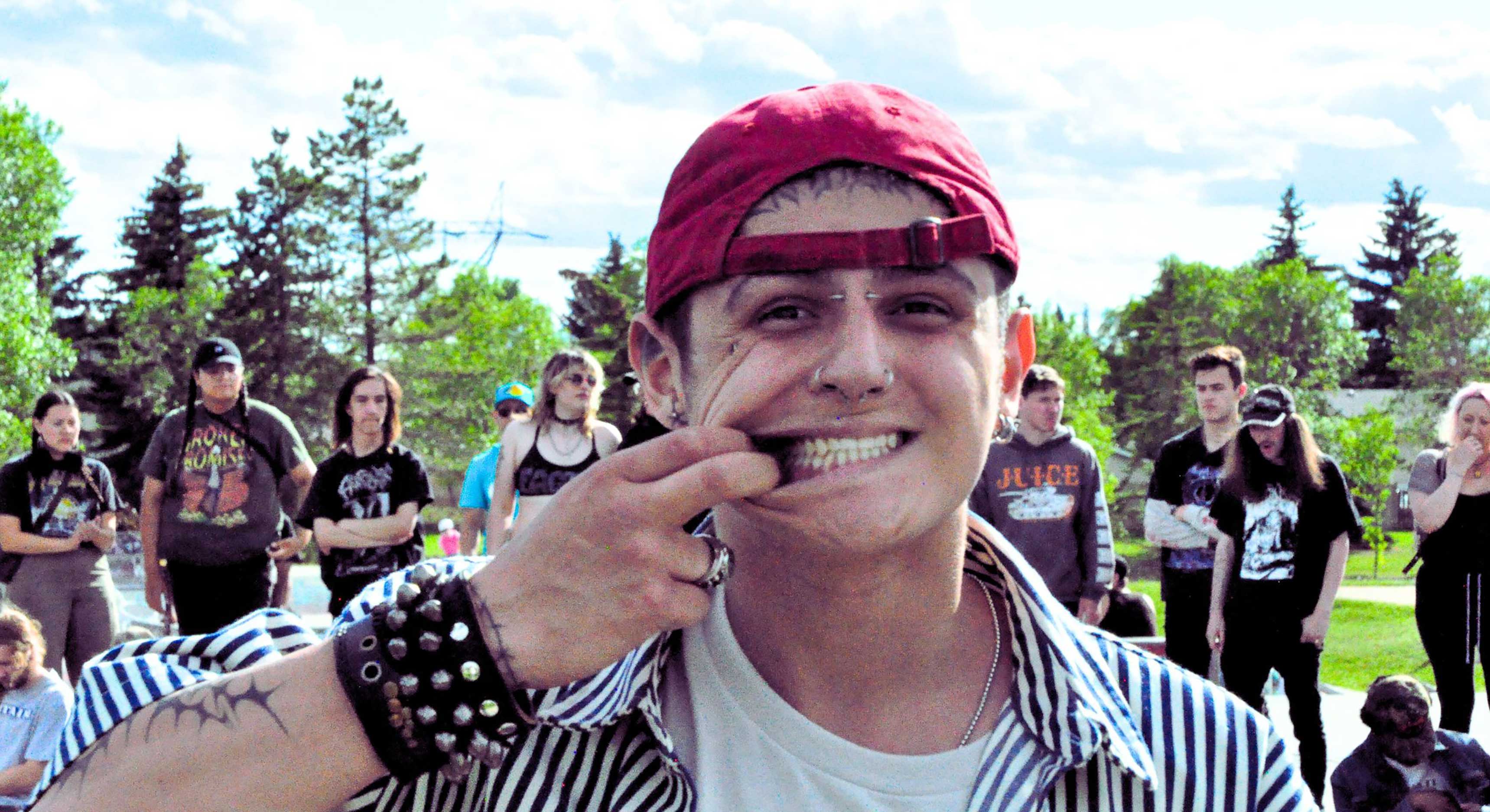 Kaden is wearing a backwards red ball cap and a striped black and white button-down shirt over a white T-shirt. He has several facial piercings and is smiling at the camera, stretching his smile with one hand. Behind him are young people standing in a skate park. 
