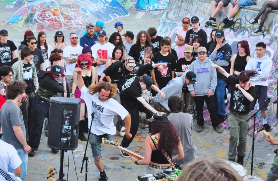 Musicians are playing while people move to the music in the bowl of a skate park. Behind them are skate park jumps and walls covered in graffiti.