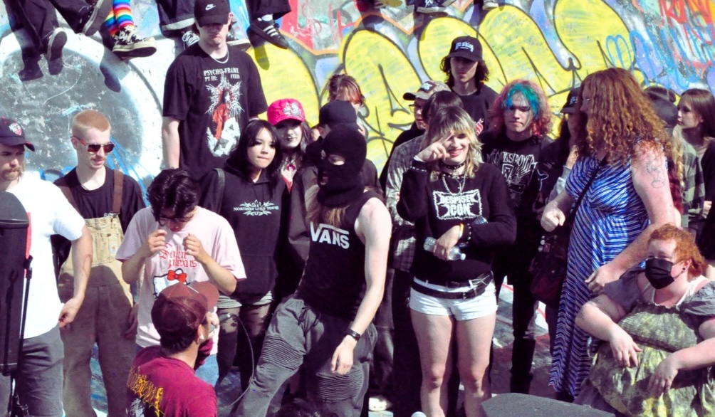 A group of young adults in casual streetwear gather in the bowl of a skate park for the Wildrose Castle Jam. They are standing outdoors against a sloped concrete wall covered in colourful graffiti.