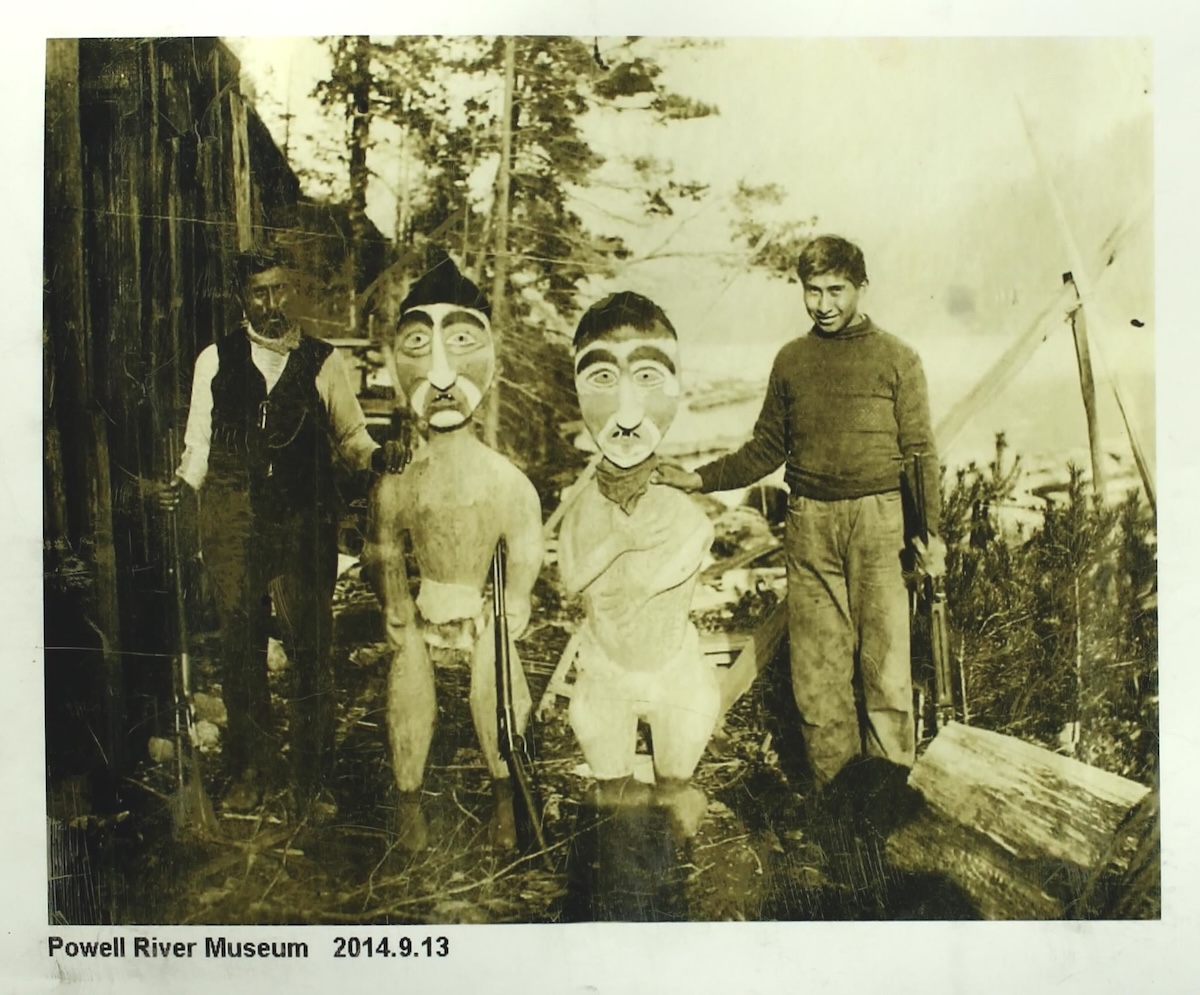 An archival black and white photo features two people standing next to two mortuary poles, carved wooden figures that are almost as tall as those next to them.