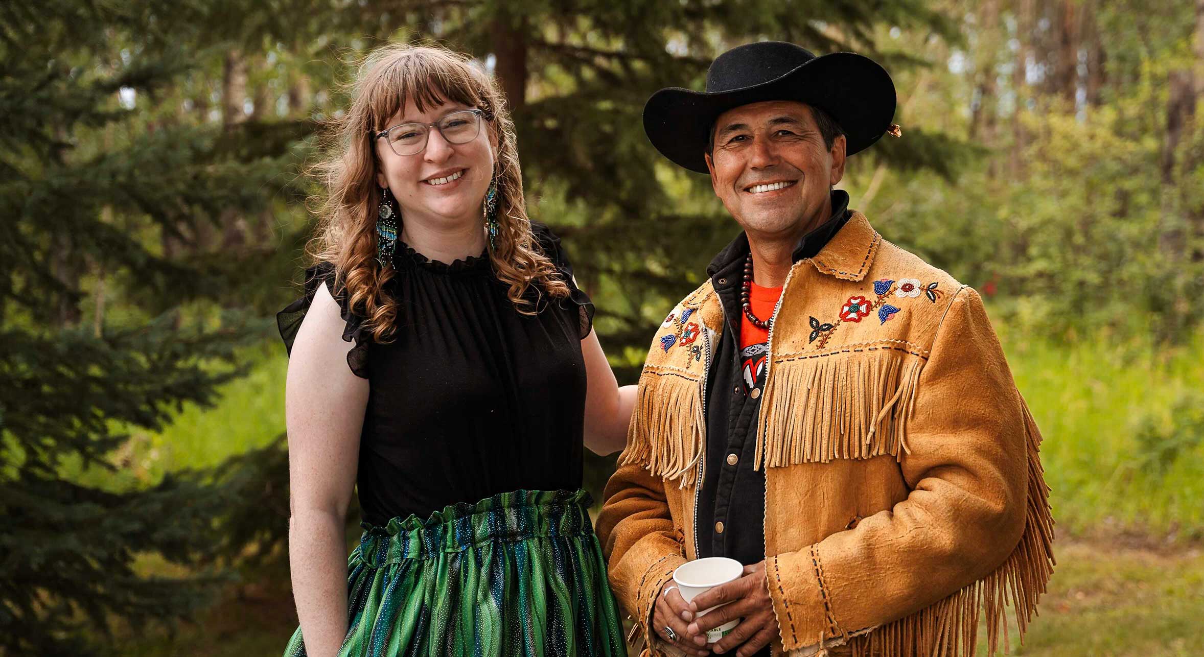 Alyssa Currie, left, stands with Elder Garry Oker, right, against a stand of evergreen trees. Currie has long, wavy light brown hair and glasses. She is wearing a ruffled black top and long colourful green skirt. Oker is wearing a black cowboy hat and a light brown leather jacket with embroidered flowers over a black top and orange T-shirt.