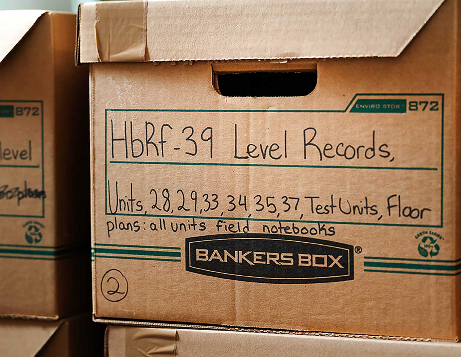 Top: three people stand together near a shelf stocked with cardboard boxes, gesturing and smiling in celebration. Bottom: a close-up of a bankers&rsquo; box with its contents labelled in handwriting using a black marker.