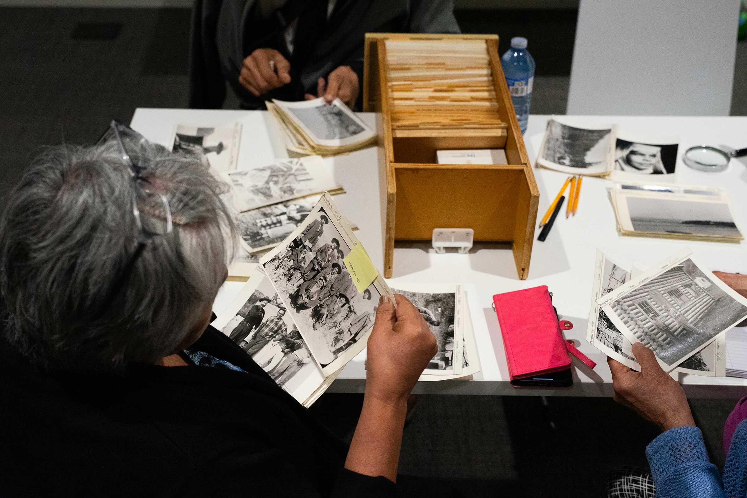 A bird&rsquo;s-eye view of people working together at a table features Virginia Gagnon, a woman with short grey hair and glasses, looking at black-and-white archival photographs of the Cheslatta Carrier Nation. The photos are assembled across the table, upon which sits a narrow wooden tray of cards.