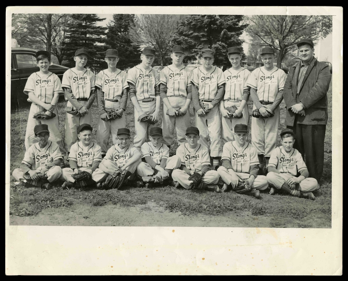 A black and white photograph of a teenage boys&rsquo; baseball team posing for a group photo, the back row standing, the front row sitting on the grass. The boys wear caps and baseball gloves and have Stong&rsquo;s written on their striped shirts. All are white except for Howard, who sits far left; the coach stands far right.