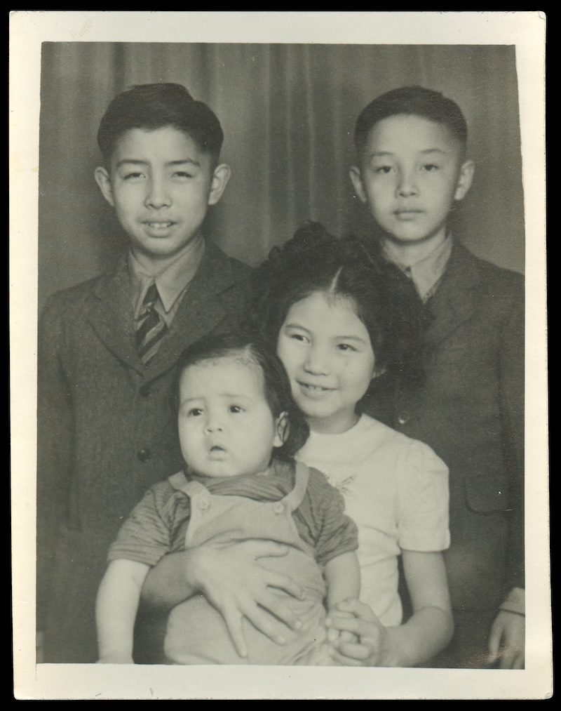 A black and white studio portrait of four children seated and standing against a curtained backdrop. Larry and Gordon are wearing suits and standing at the back. Helen is wearing a white shirt and smiling with Howard, in a jumper, on her lap.
