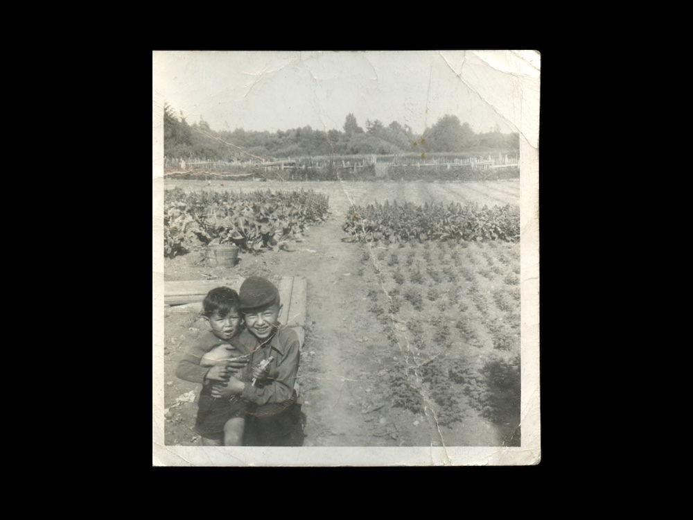 A crinkled black and white photo features two young boys at the lower left bottom of the frame. Larry Grant, right, stands holding Howard Grant, left. Larry smiles as he cuddles his brother, who has his mouth open in surprise. Rows of vegetables are planted to the left and behind them.