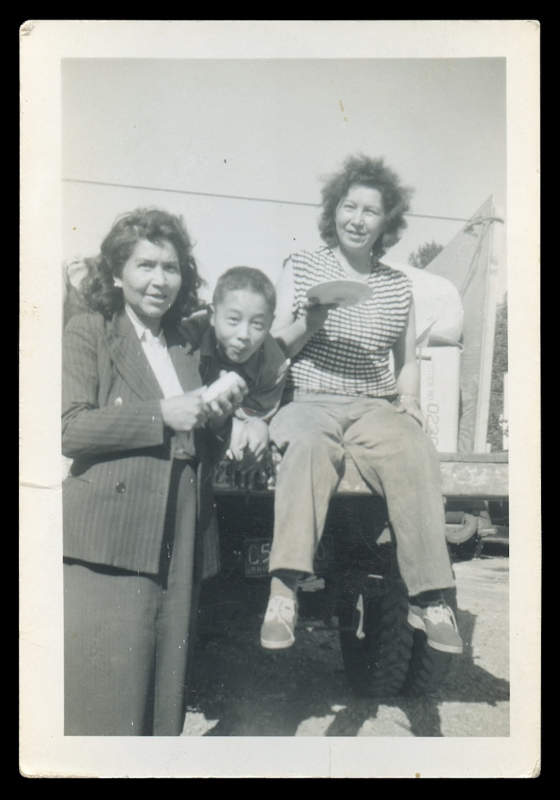 A black and white photo depicts two adult women standing or seated on either side of a school-aged boy, who sticks his head between them, a puckish expression on his face. They are posing on the back of a pickup truck on a sunny day.