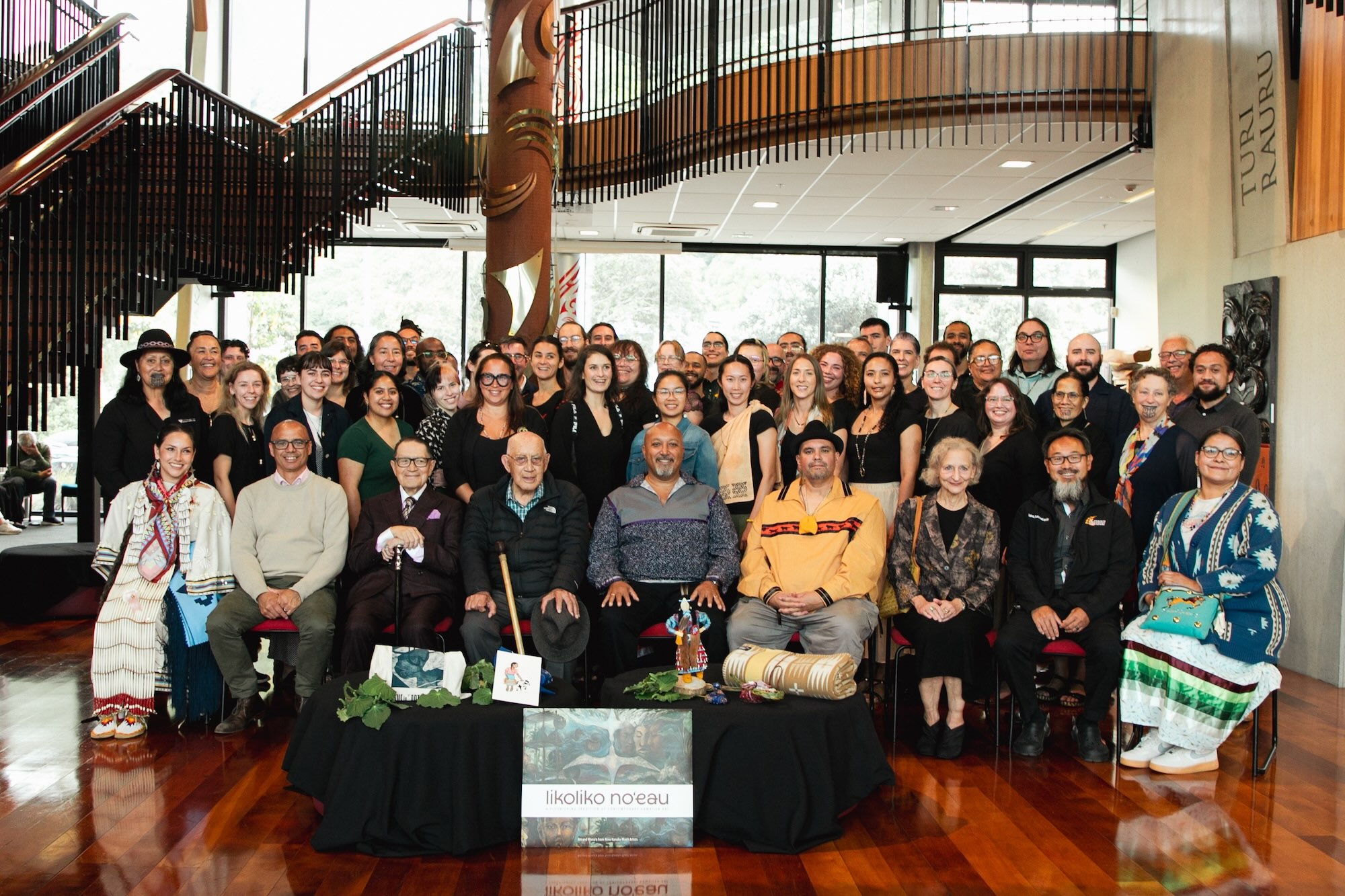 A large group of people who comprise the Abundant Intelligences Research Program pose for a photo in an indoor space. They are standing and seated behind a low black table festooned with Indigenous ceremonial offerings including plants, a blanket and artwork. 