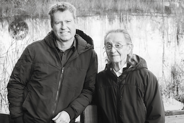 A black and white photograph of Scott Steedman and Larry Grant standing outdoors with a body of water behind them. They are smiling at the camera and wearing dark hooded jackets.