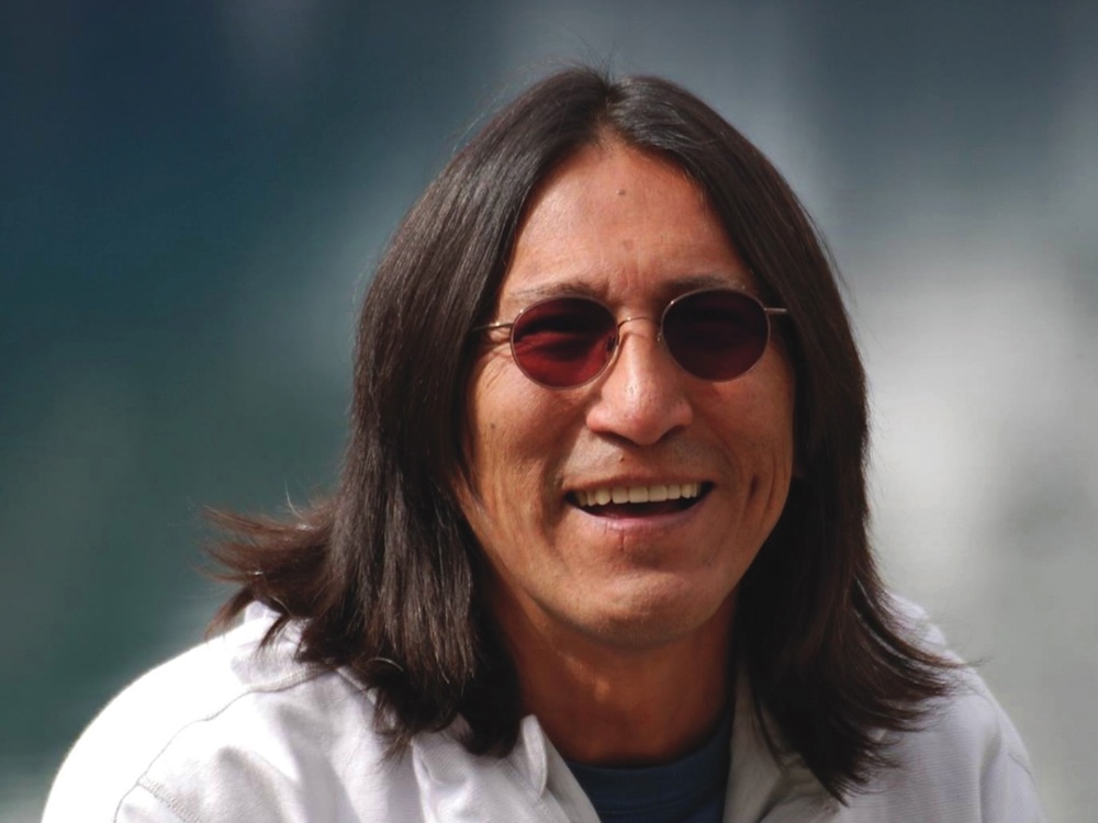 Richard Wagamese has shoulder-length dark hair and a medium skin tone. He is wearing dark glasses and a white collared shirt over a blue T-shirt. Blue and white tones are in soft focus behind him. He is smiling.