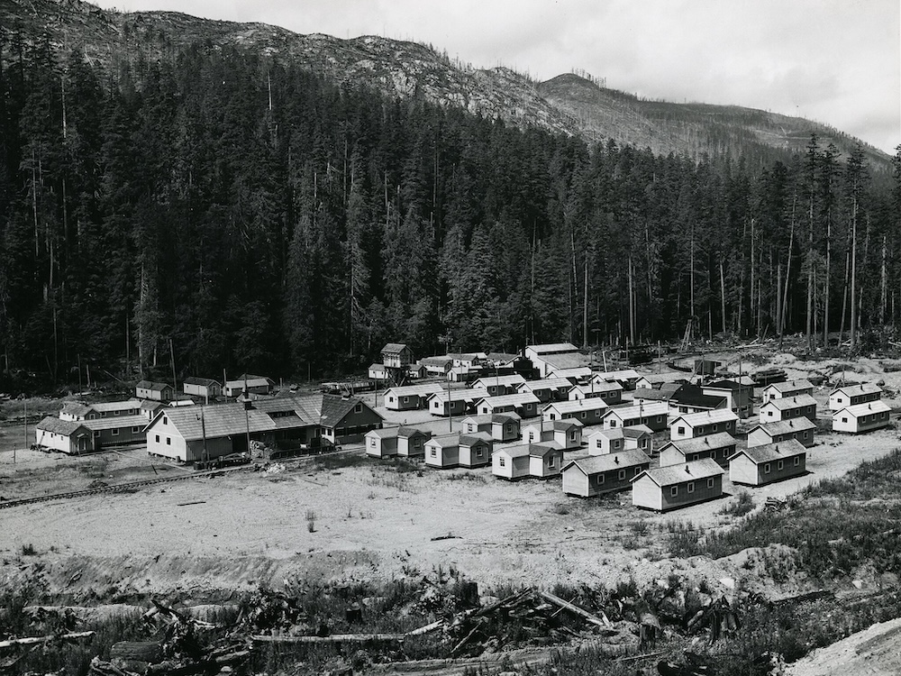 A black and white photo shows a worker camp featuring more than a dozen small modular homes along with several larger buildings.