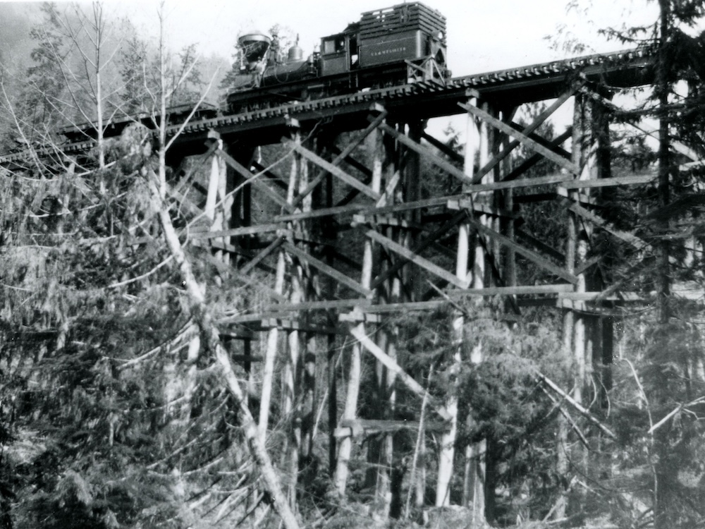 A black-and-white image of a steam train crossing a high wooden train trestle above the forest floor.