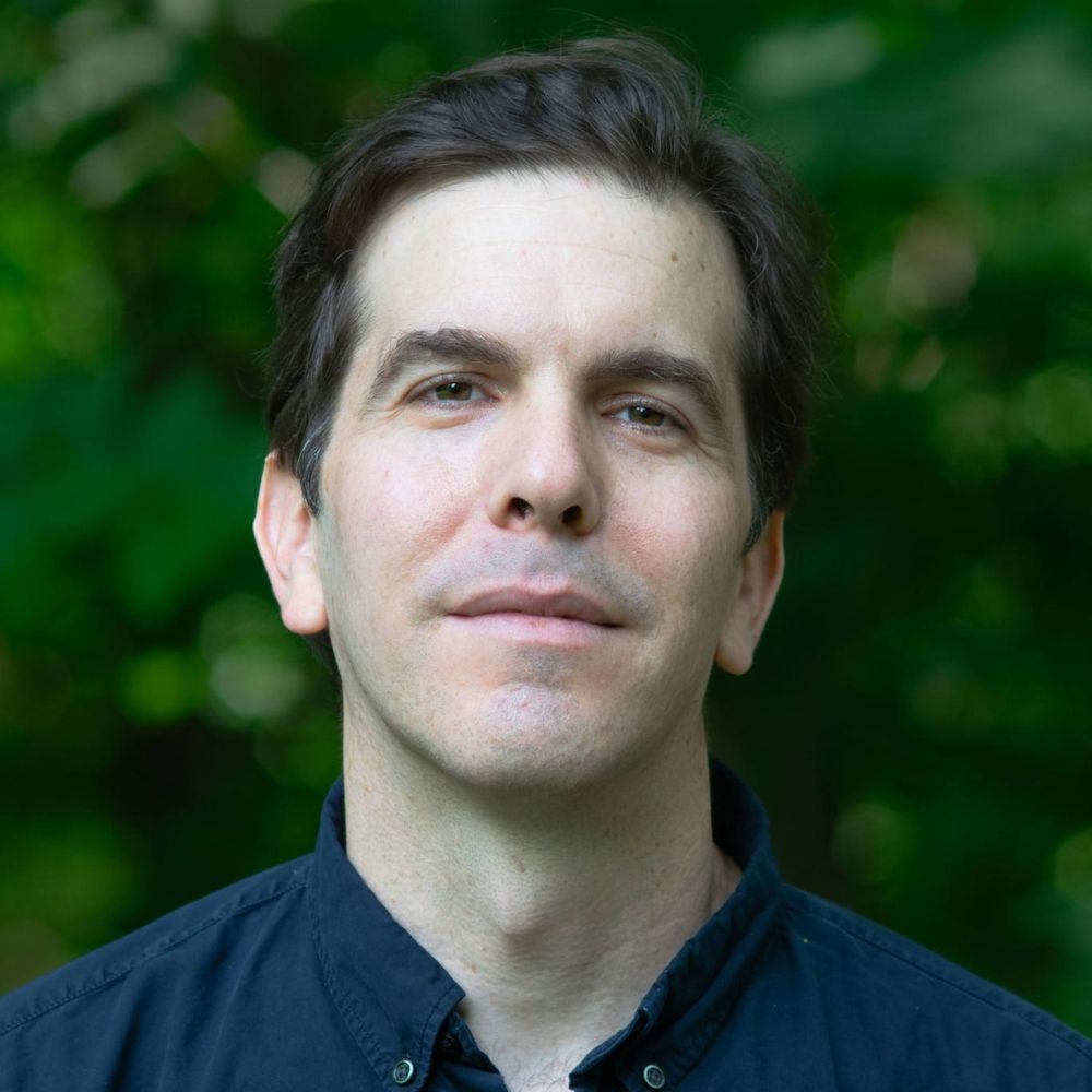 Jacob Silverman has dark brown wavy hair and a light skin tone. He is wearing a navy collared shirt and standing against a soft-focus green background outdoors.