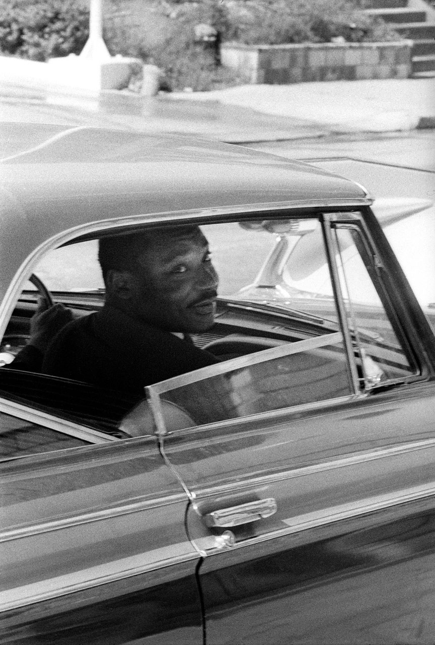 A black and white documentary-style photograph depicts the right-side profile of Martin Luther King Jr., a Black man in a dark suit, looking gently to the side. He is in the passenger seat of a car.
