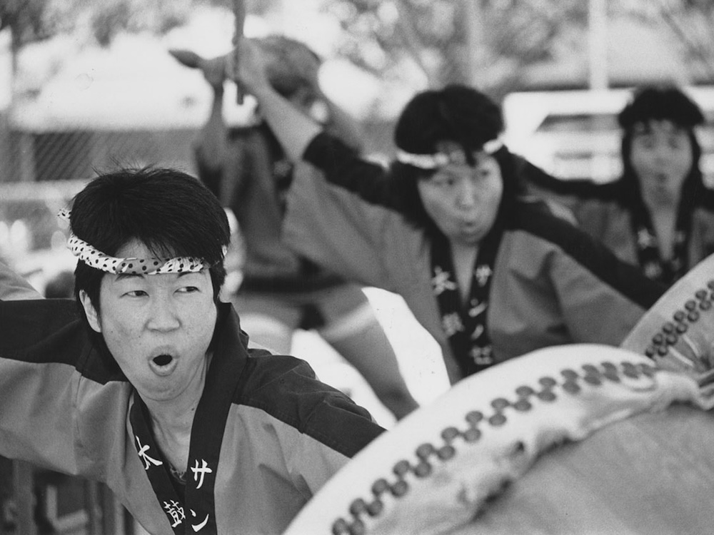 A black and white photograph depicts three women playing the taiko drums at an outdoor festival. They all have short black hair and are wearing headbands and traditional Japanese regalia.