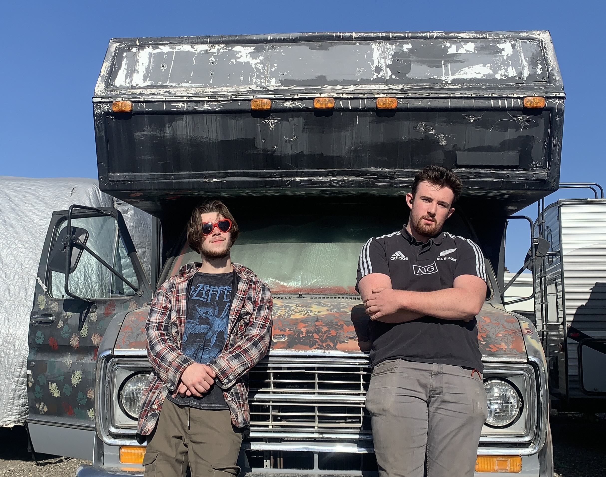 Two young men stand in front of a recreational vehicle covered in black and red paint on a sunny day. Adam Sweet, left, has wavy brown hair and is wearing red heart-shaped sunglasses. Thomas Cormier, right, has short brown hair and is wearing a black Adidas T-shirt.