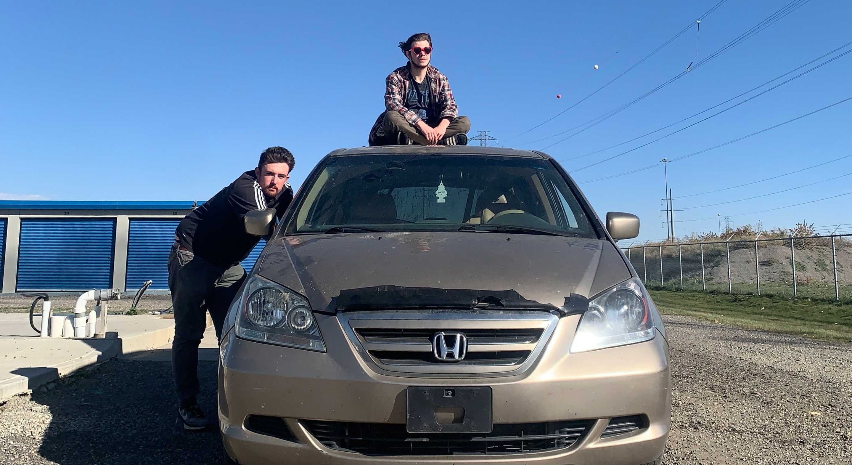 Two young men pose with a worn champagne-coloured Honda van. Adam Sweet sits atop the roof and Thomas Cormier leans against the passenger window. The car is parked on a gravel roadway under an open blue sky on a bright, clear day.