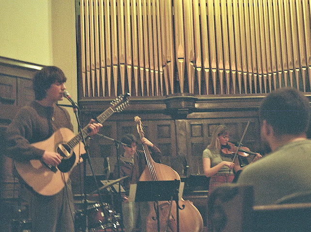 Sawyer Begg stands at a microphone against a background of gold organ pipes in a church. He has wavy brown hair and is holding an acoustic guitar. A cellist and a violinist are playing to the right of the frame.