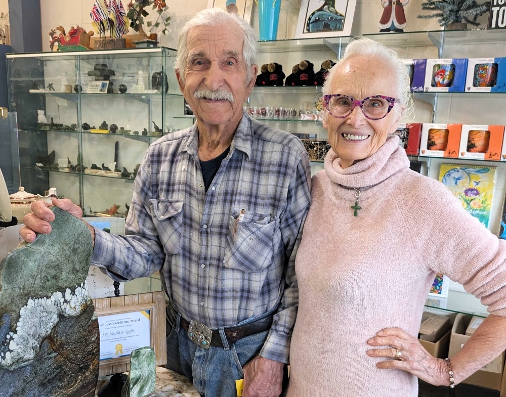 An elderly light-skinned man with white hair and a bushy moustache smiles at the camera. Beside him, an elderly woman with white hair stands with one hand on her hip. Both people stand behind the counter of a gift shop.