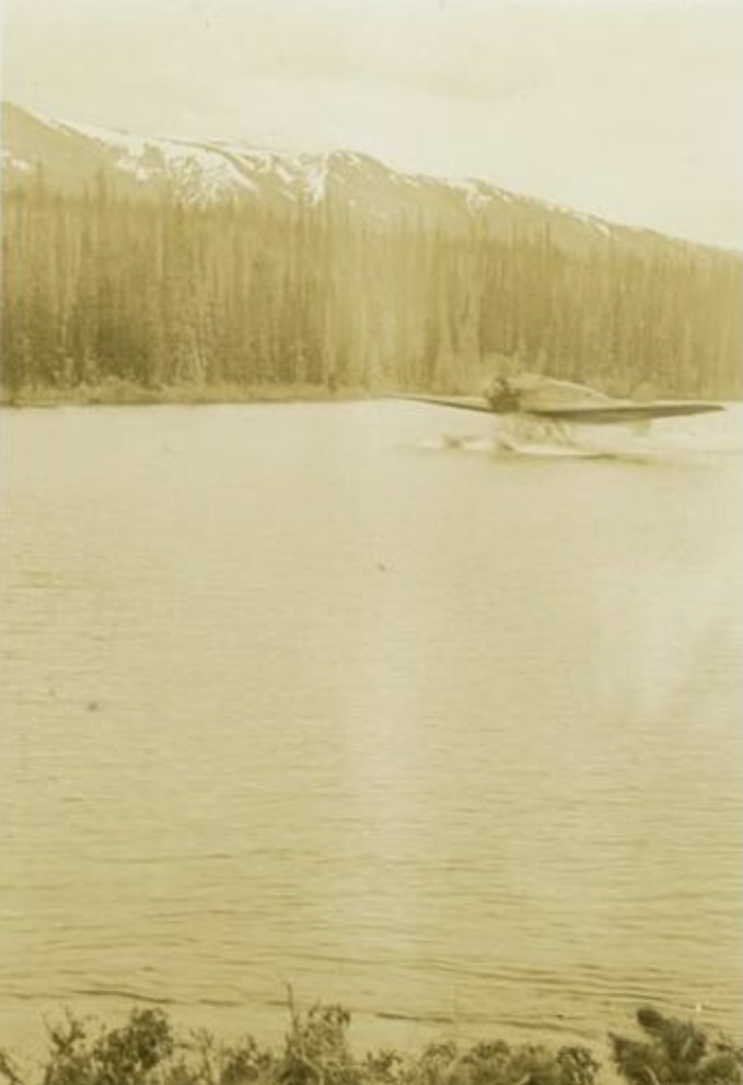 A sepia-toned archival photo of a float plane on a northern lake with trees and mountains in the background.