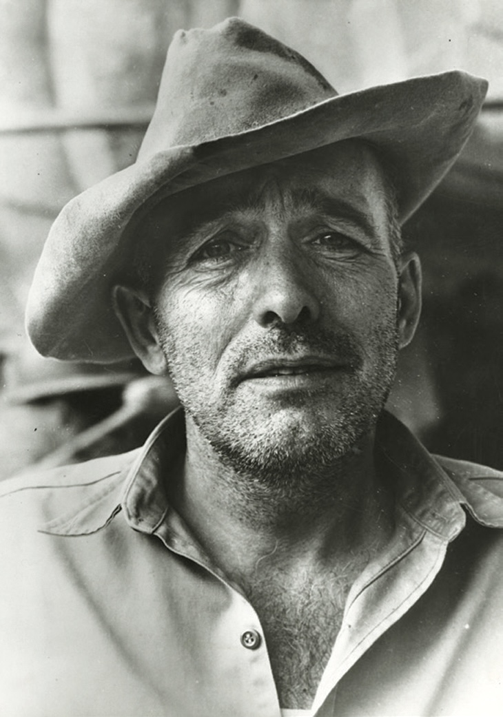 A black and white photograph shows a light-skinned man&rsquo;s lined face with stubble, topped with a battered western-style hat worn on a rakish angle. He wears a button-down shirt open at the collar.