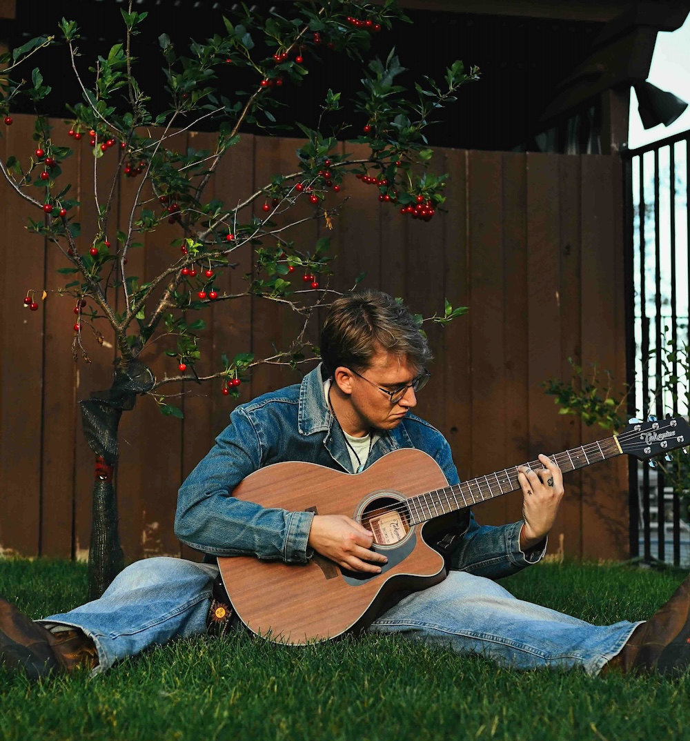 Stephen Curtis is seated in the grass in a yard against a brown shed, under a small tree growing red berries. He has short blond hair and is wearing a blue jean jacket and blue jeans, playing acoustic guitar.