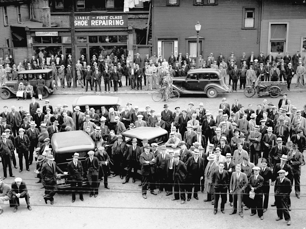 An old black and white photo, taken from above, of a crowd of men, nearly all dressed in suits and fedoras, looking up in the direction of the camera. Some are leaning on 1930s-era automobiles parked among the crowd.