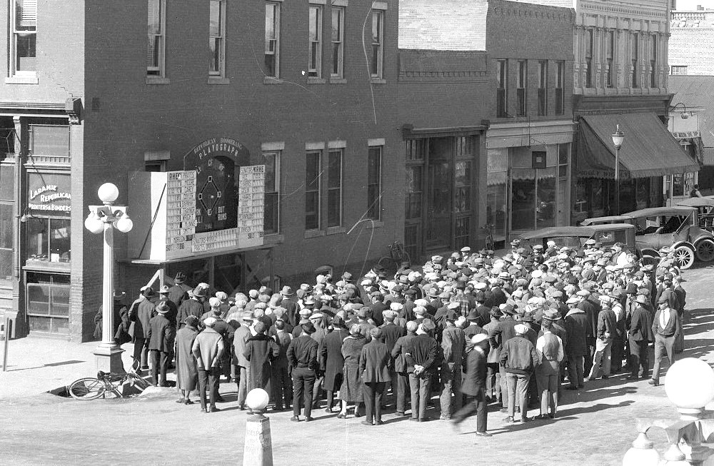 An old black and white photo of a crowd of people in 1920s attire looking up at a Playograph on the side of a brick building. 
