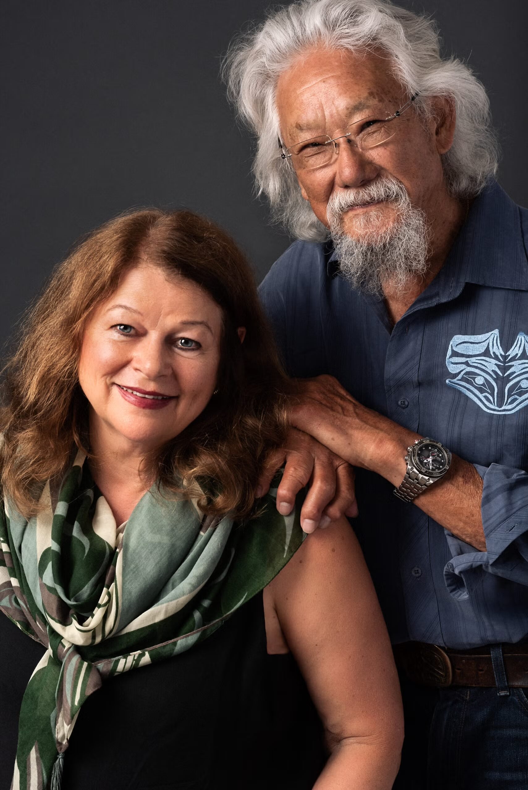 Tara Cullis, left, has wavy red hair and is wearing a green patterned scarf and a black top. David Suzuki, right, has wavy white hair and glasses, and he is wearing a blue-grey buttond-down shirt with light blue embroidery. They are posing against a grey studio background.