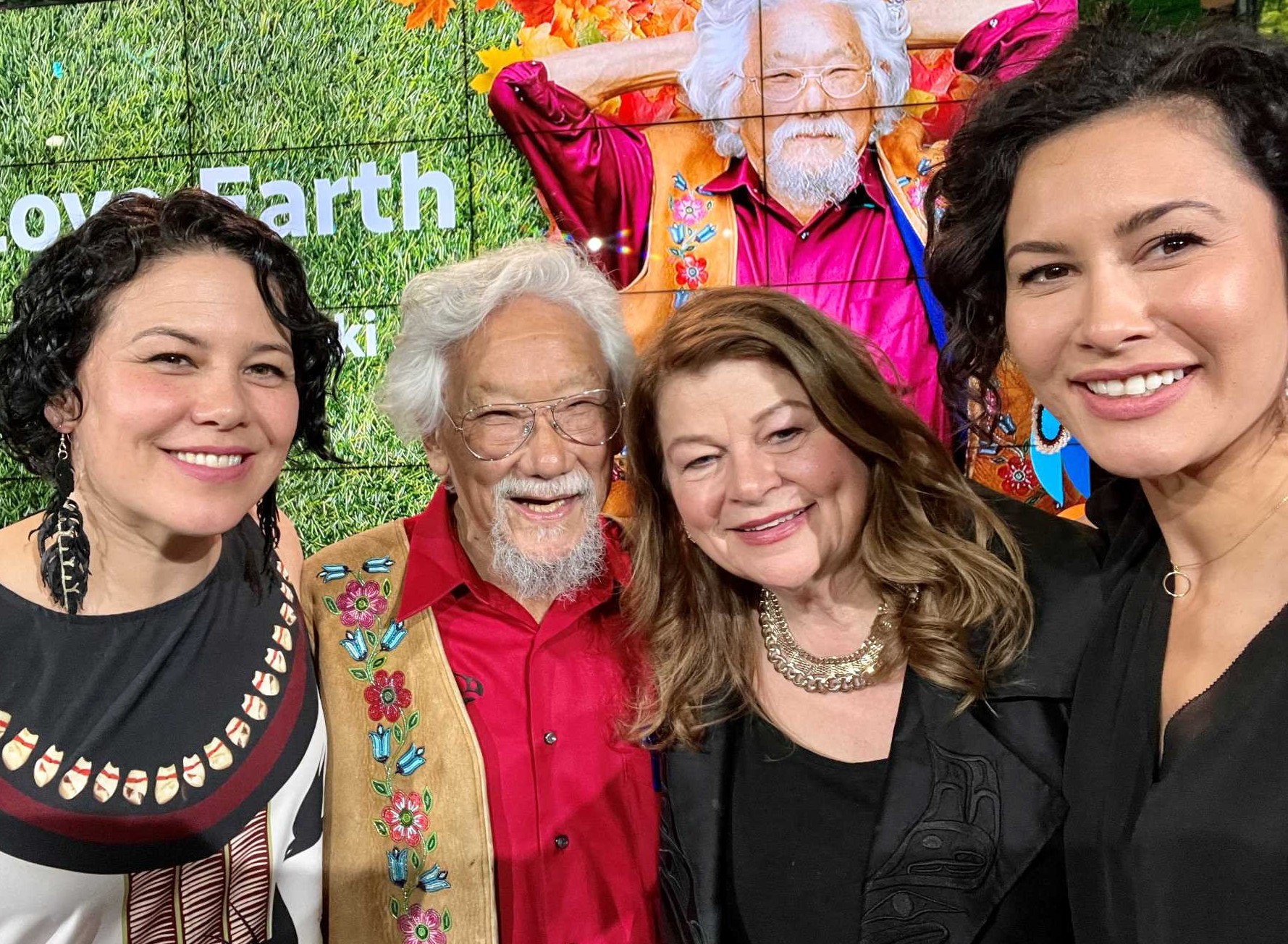 Four people pose for a photo against a digital background featuring a photo of David Suzuki lying in the grass. From left to right, Severn Cullis-Suzuki, David Suzuki, Tara Cullis and Sarika Cullis-Suzuki pose for a photo at a party. They are smiling at the camera. 