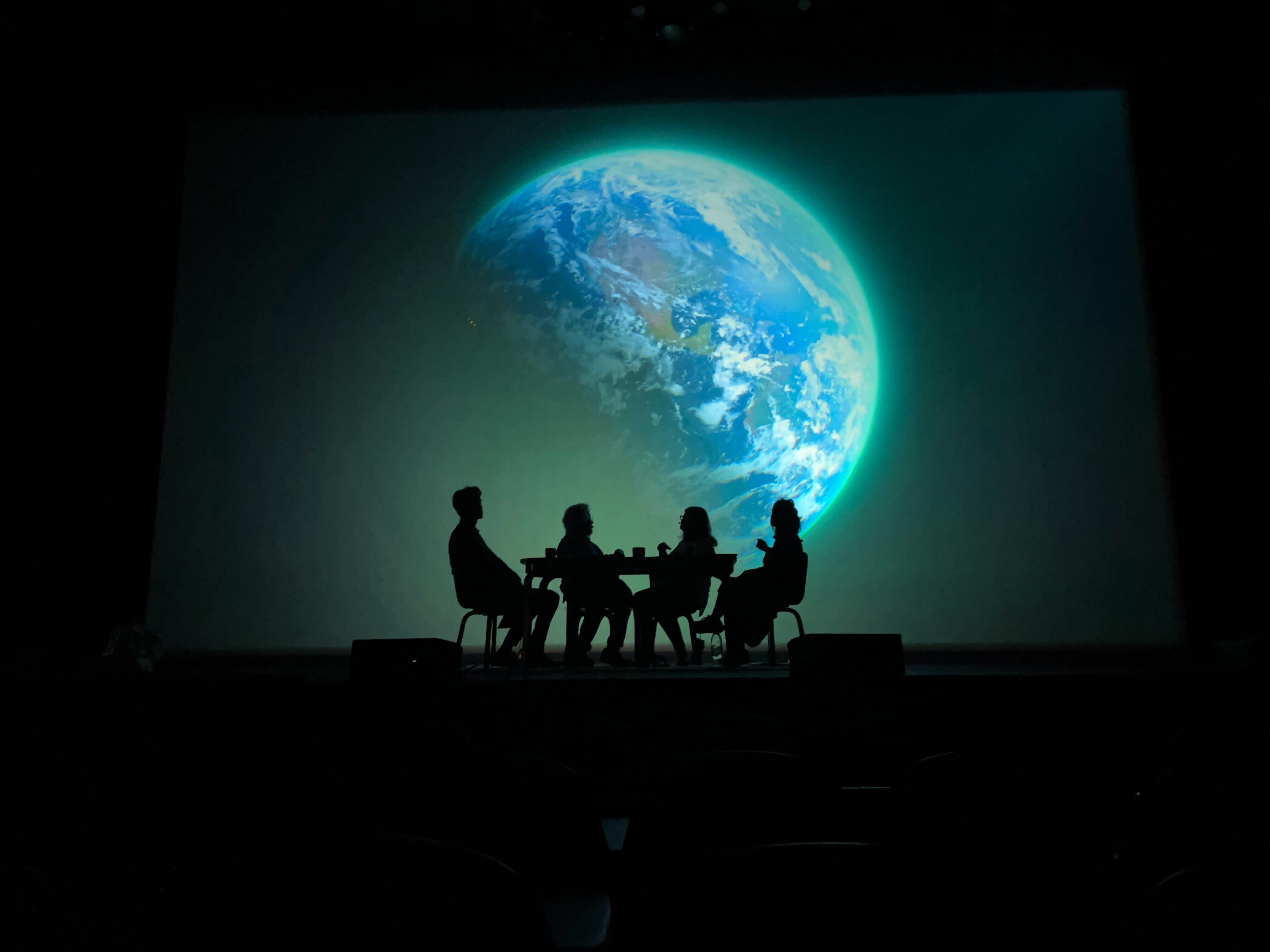 Four people in silhouette sit at a table on a darkened stage. Behind them is a large projected image of the planet Earth ringed with green light.