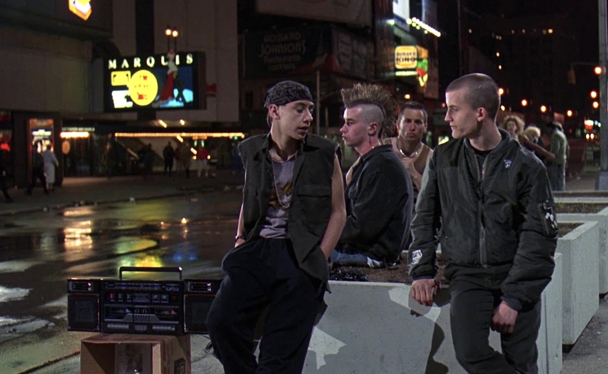 Four young men lean against a set of white concrete barriers along a downtown street at night. They are clad in casual streetwear reminiscent of the late 1980s. A boom box sits on a small wooden box to the left of the frame.