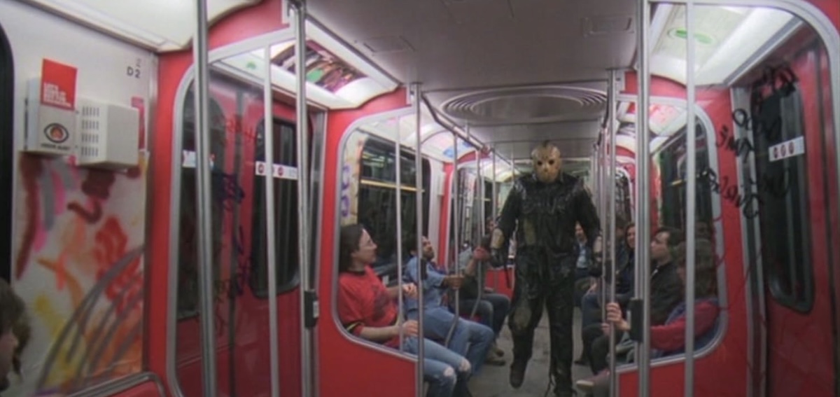 A man wearing a dark outfit and a hockey mask walks down the middle aisle of a subway car with red doors. Passengers are seated on either side of him, some of whom are gripping the train&rsquo;s poles.