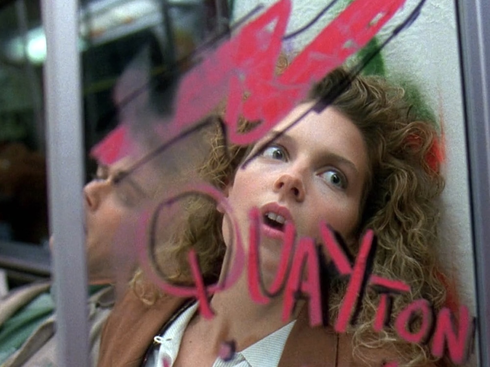 Actor Jensen Daggett has long curly strawberry-blond hair and a light skin tone. She looks through the graffitied glass partition of a subway train with horror. There is a man in a green shirt in the seat beside her and his face is obfuscated by the glass and a pole on the train.