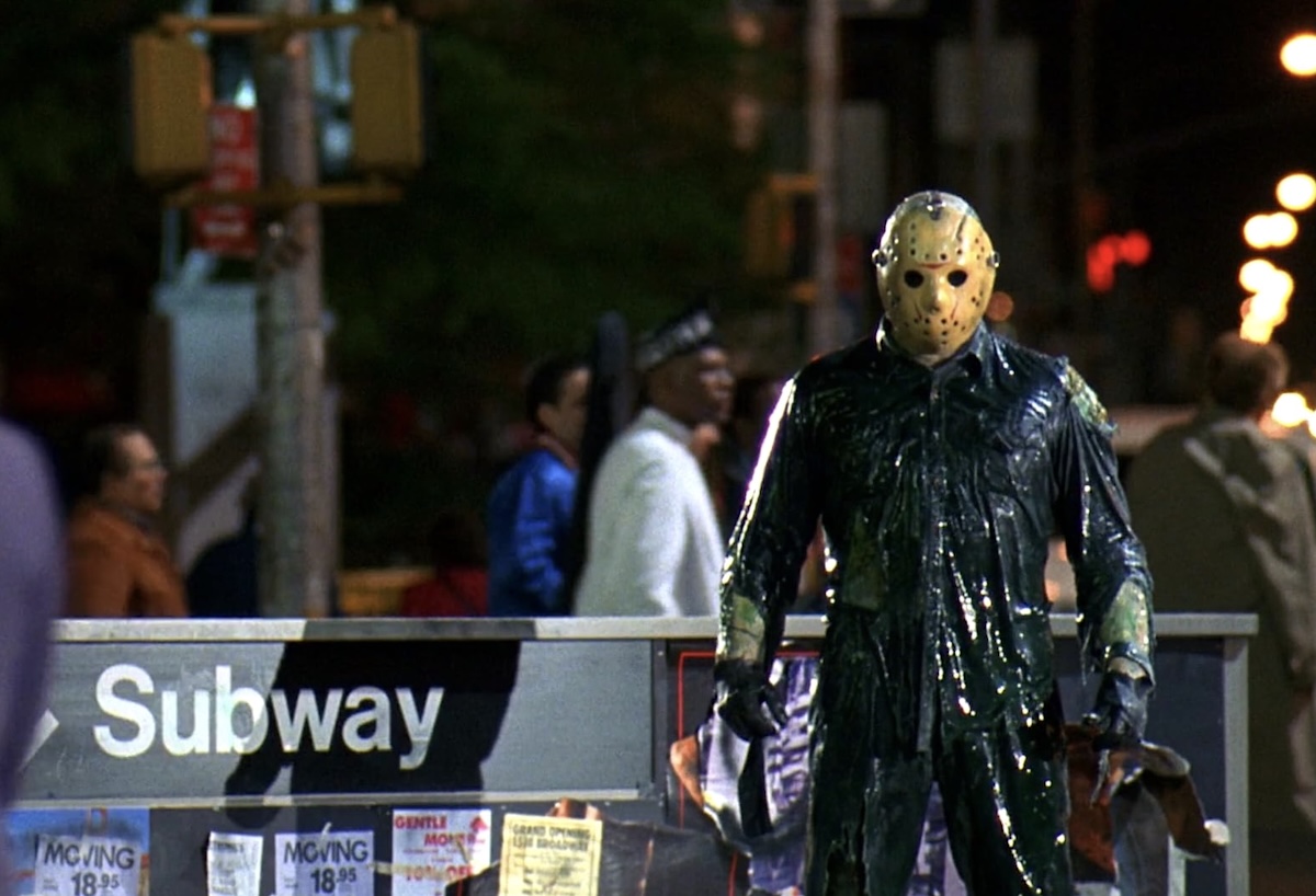 A figure in a black plastic suit and a hockey mask stands on a downtown street at night. To his left is a low black sign that reads &lsquo;Subway.&rsquo;