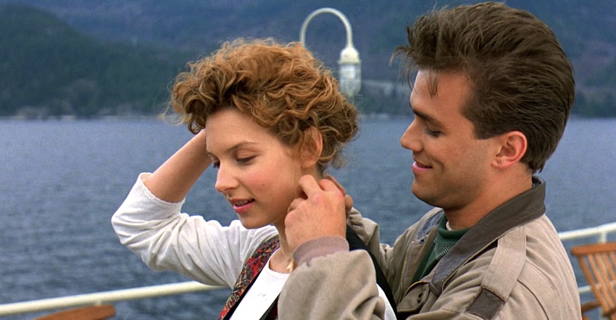 Actors Jensen Daggett, left, and Scott Reeves, right, interact on the deck of a boat with white rails. Behind them is a mountainous landscape and blue water.