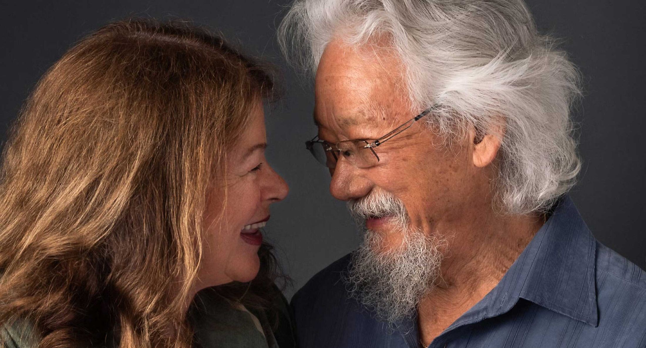 A studio portrait of David Suzuki, right, with Tara Cullis, left. The couple is looking at each other and smiling. Cullis has wavy red hair and a pale skin tone. She is wearing a sage green shirt. Suzuki has wavy white hair, glasses, a beard and a medium skin tone. He is wearing a blue-grey shirt with light blue embroidery. They are standing against a dark grey studio background.