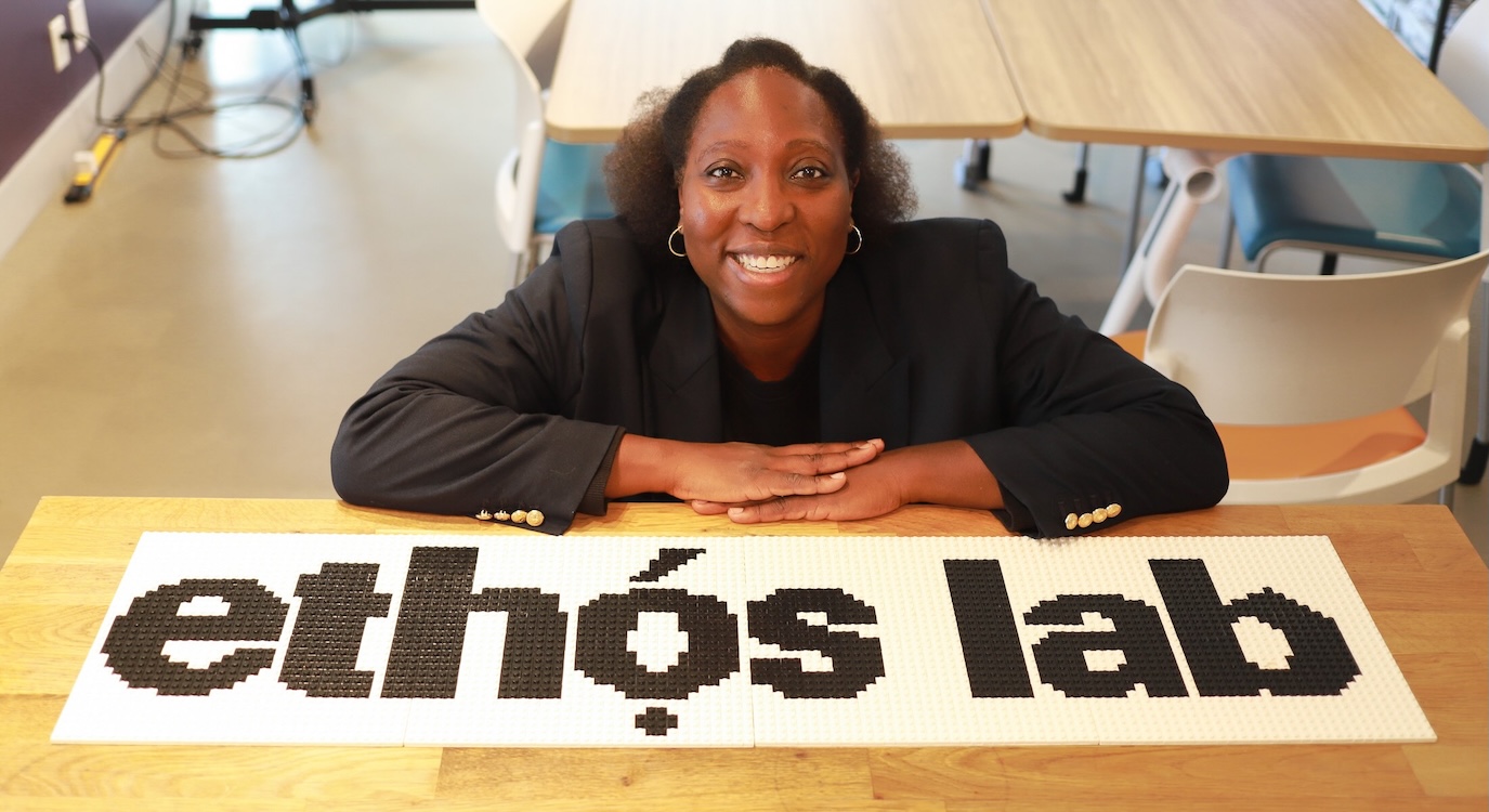 Anthonia Ogundele is seated in a classroom-style laboratory. She is a Black woman with short wavy dark hair and a black blazer. The words &ldquo;Ethos Lab&rdquo; are printed in black across a white Lego grid.