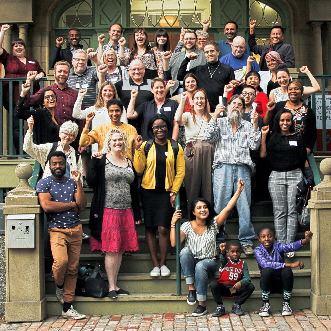 A group of people stands and sits on the front steps of a building, raising their fists in solidarity and smiling.
