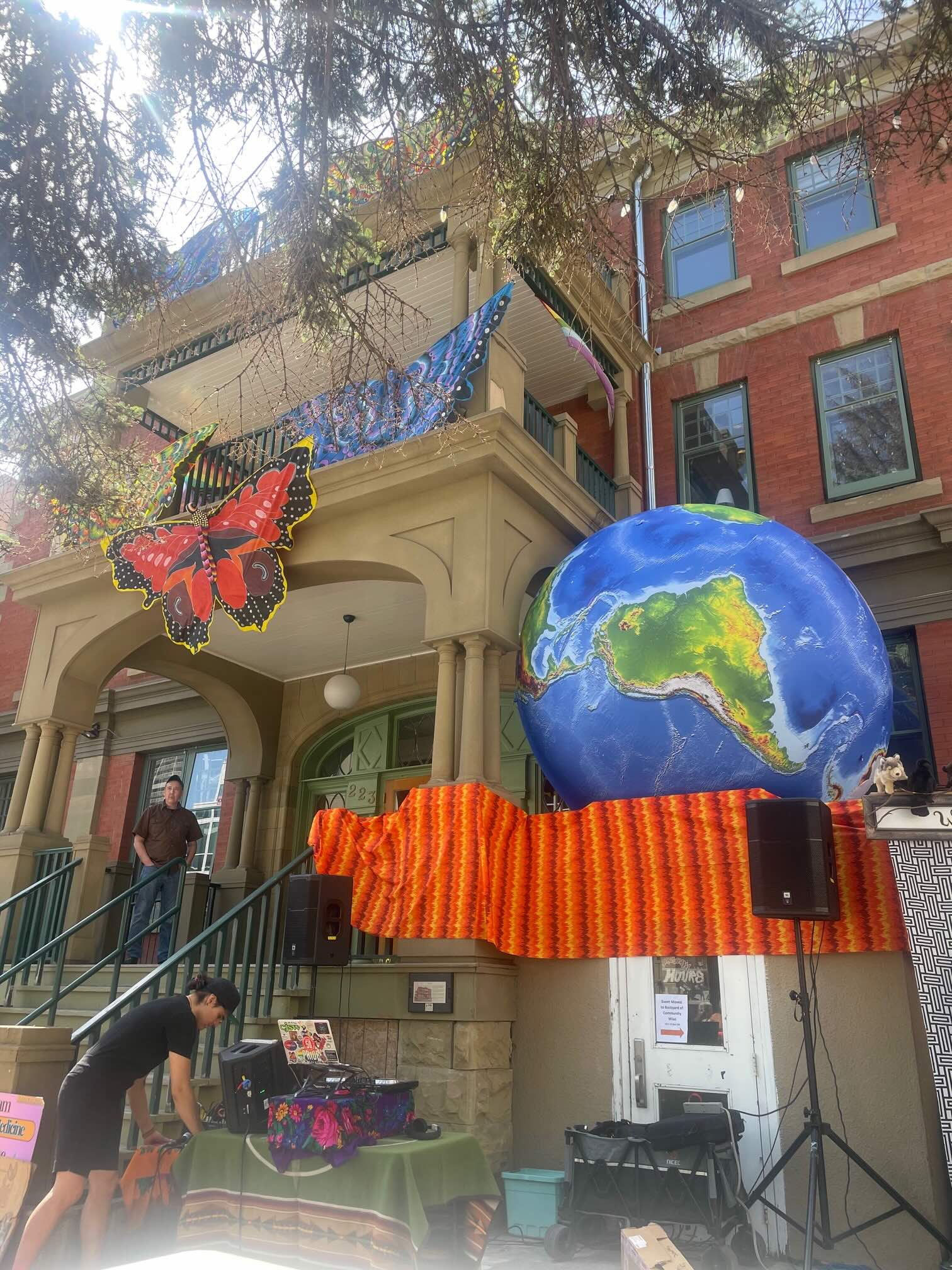 A large butterfly and planet earth installation decorate the Georgian Revival fa&ccedil;ade of an historic building while one person stands on the porch and another person sets up items on a table at the bottom of the front steps. 