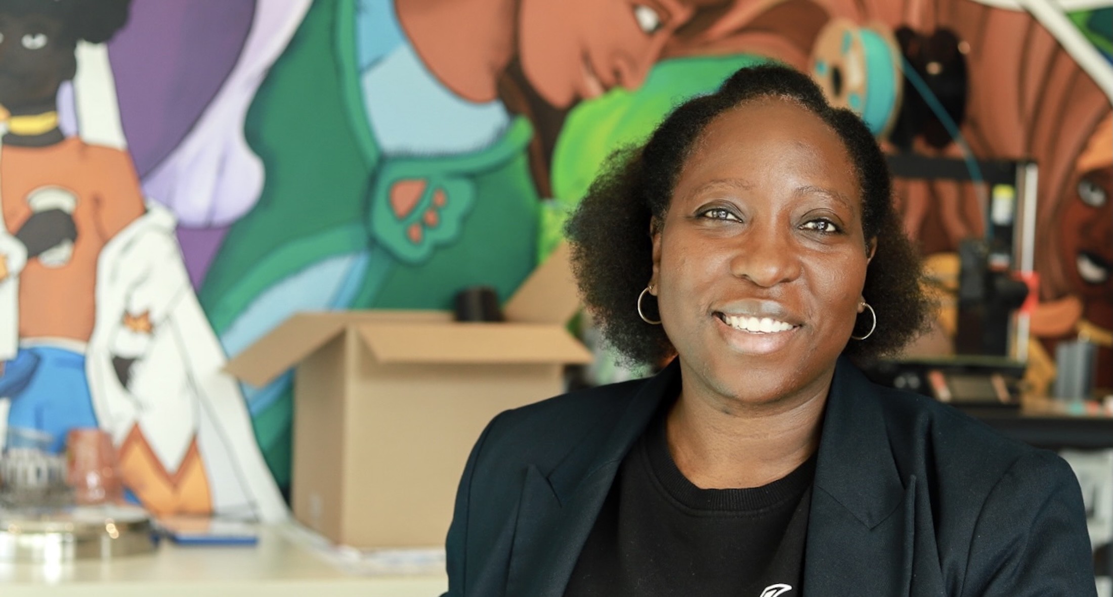 Anthonia Ogundele is a Black woman with short wavy black hair pulled back. She is wearing hoop earrings and a dark blazer over a black T-shirt. She is seated against a colourful wall painted with a mural and smiling.