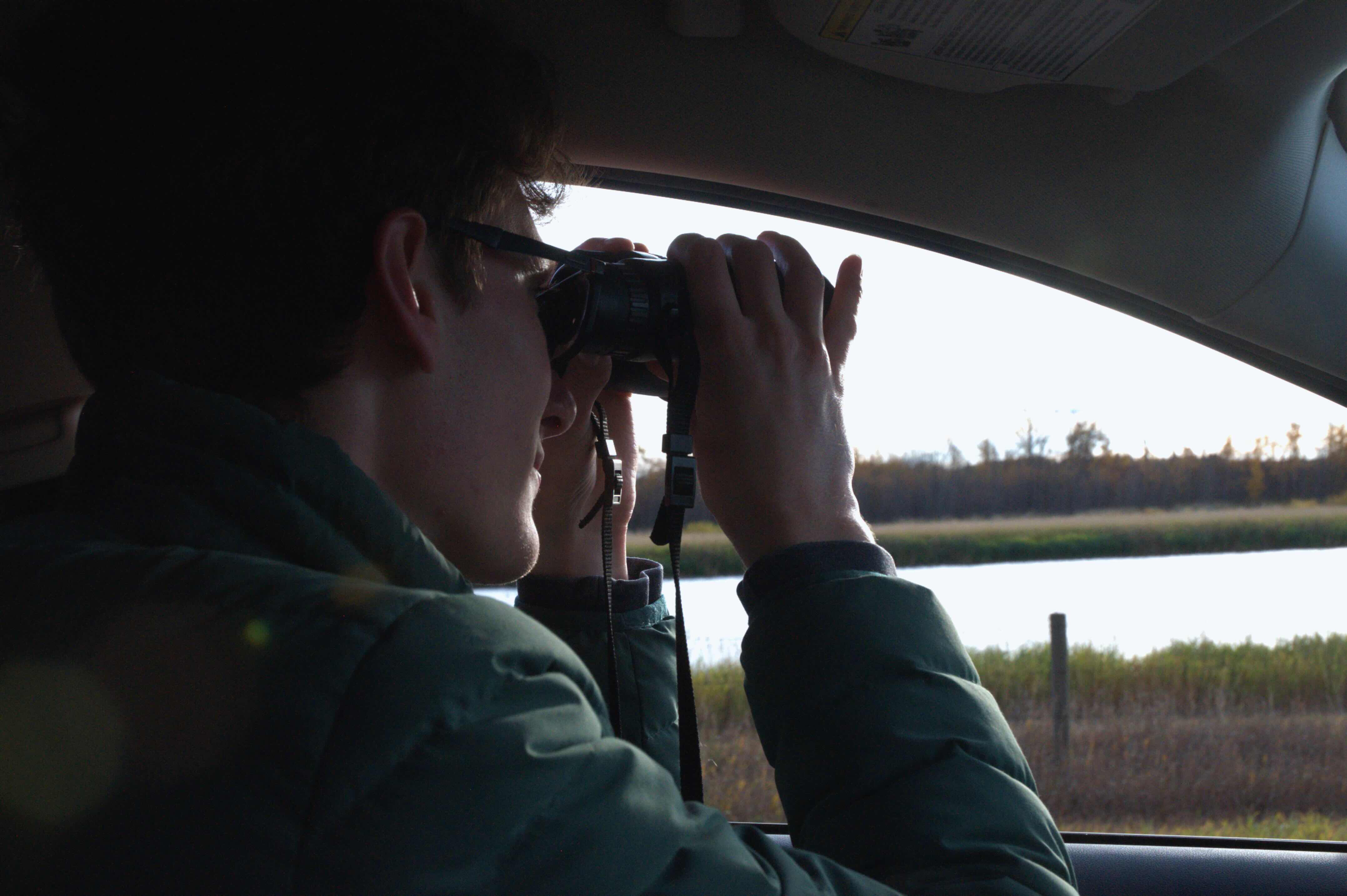 David Grinevitch is seated in the driver&rsquo;s seat of a car and looking through binoculars at a body of water outside. He is wearing a green puffer jacket and has short brown wavy hair.