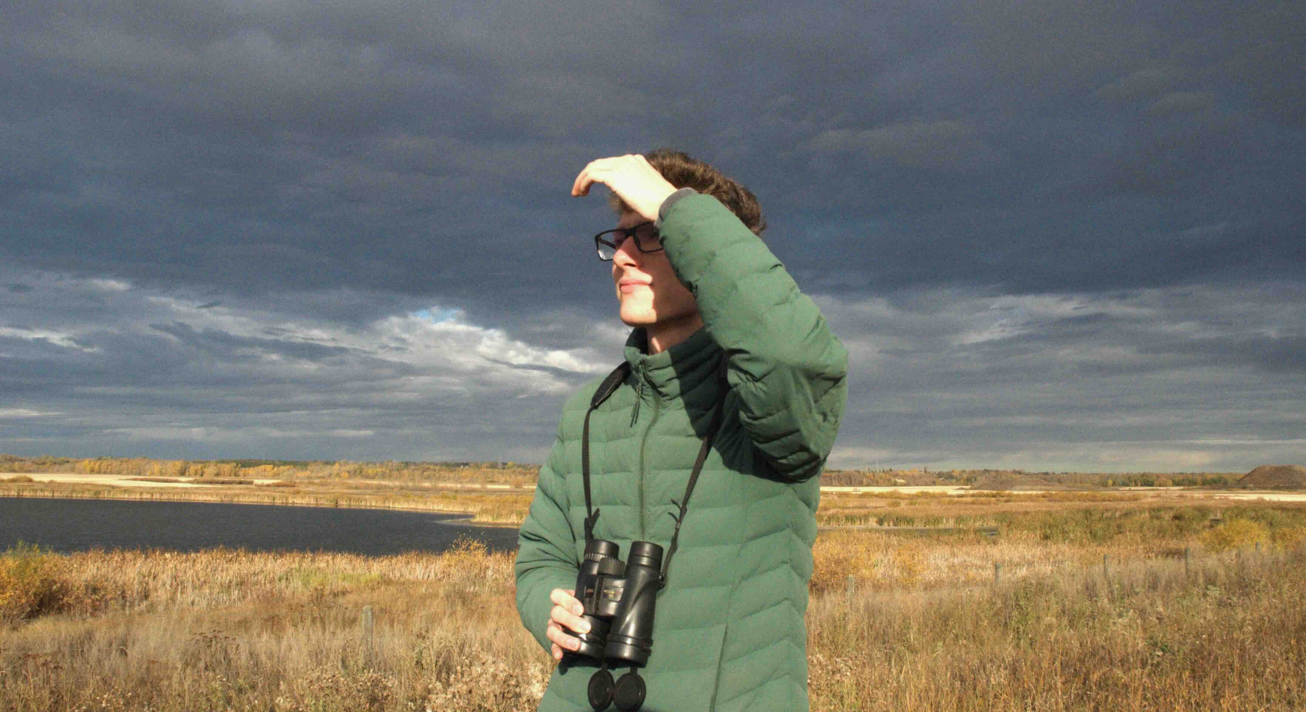 David Grinevitch is wearing a hunter-green puffer jacket and is raising one hand to his forehead to block the sun. He has short wavy brown hair, a light skin tone and glasses. He is holding a pair of binoculars around his neck. Behind him are dark grey clouds and a flat expanse of a prairie landscape.