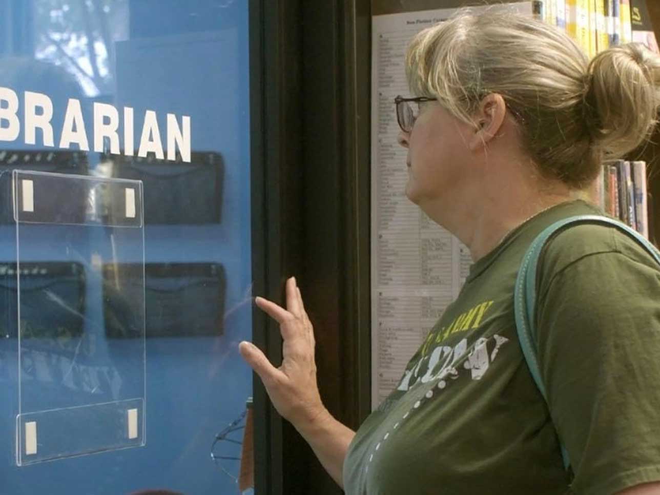 Suzette Baker is turned towards a glass door with her back towards the camera. She has blond hair tied loosely back. She has a light skin tone and she is wearing a green T-shirt and carrying a light blue shoulder bag. One hand pauses on a glass door that reads &ldquo;librarian&rdquo; in white with a blue wall in the background.