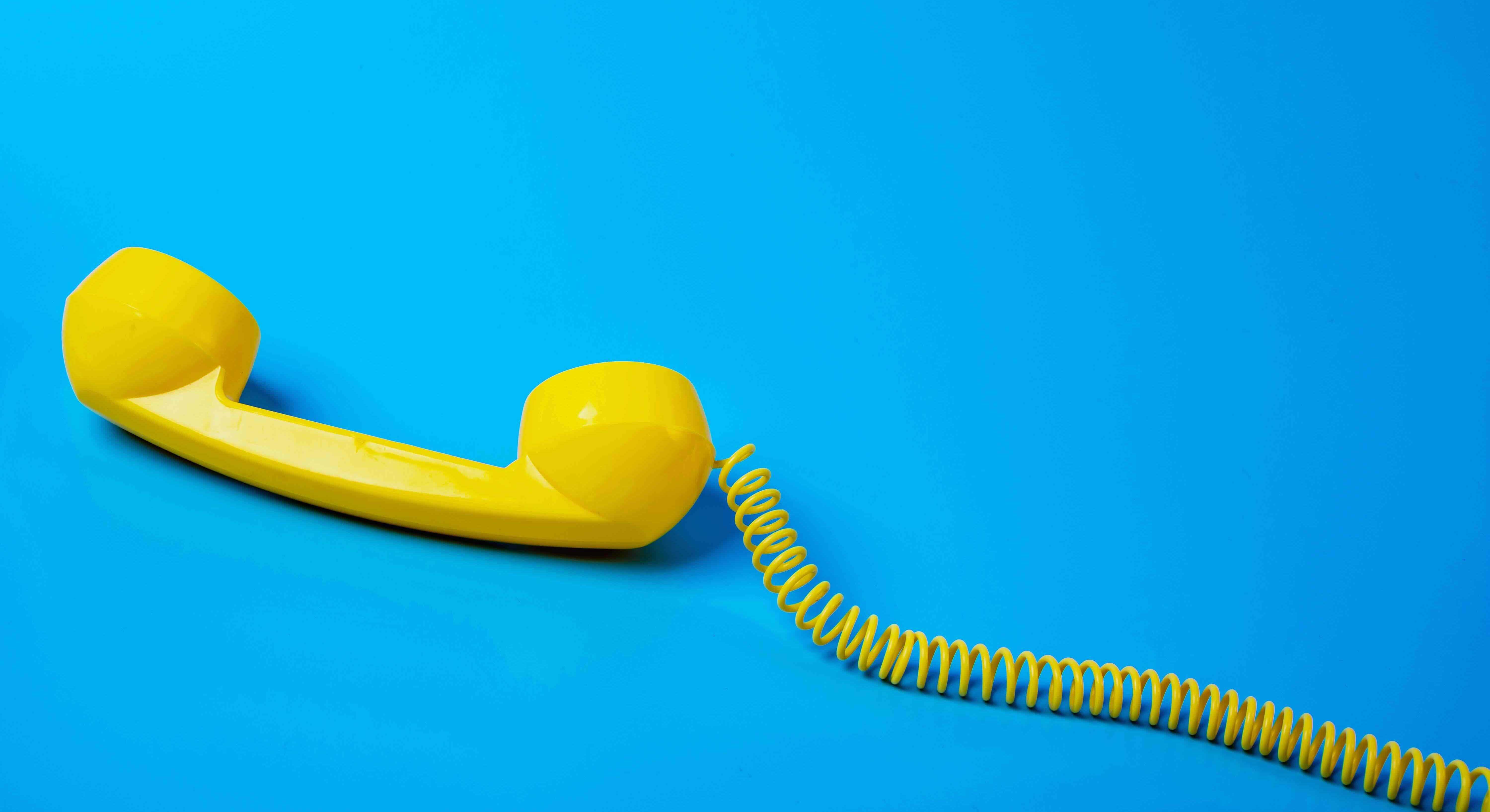 The yellow plastic receiver of a landline phone and its curly cord sits against a blue background.